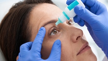 Primer plano de una persona que recibe una gota en el ojo desde un frasco dosificador sostenido por manos con guantes azules