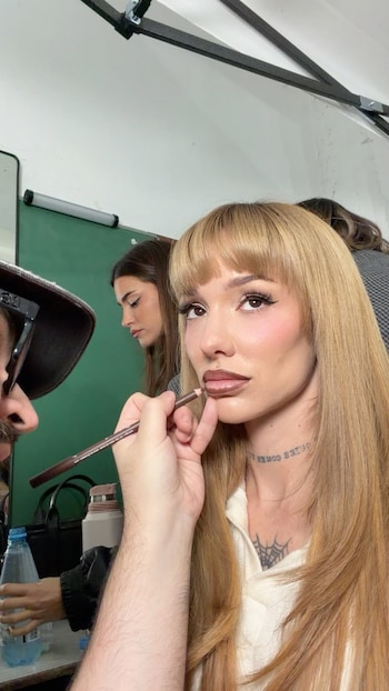 Mujer rubia con flequillo recibe maquillaje en los labios de una mano. Tiene perforación labial y tatuaje de telaraña. Otra mujer y persona con sombrero en segundo plano