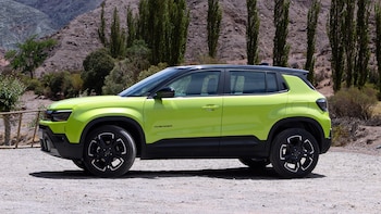 Automóvil verde con techo negro visto de perfil sobre un terreno de ripio, con árboles y una ladera montañosa de fondo