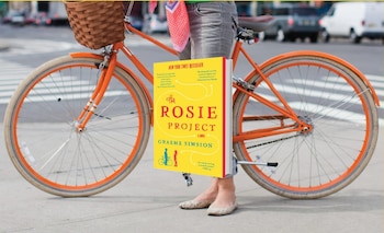 Persona con pantalones vaqueros y zapatos planos sujeta un libro amarillo con el título 'The Rosie Project'. Detrás, una bicicleta naranja con cesta y fondo urbano