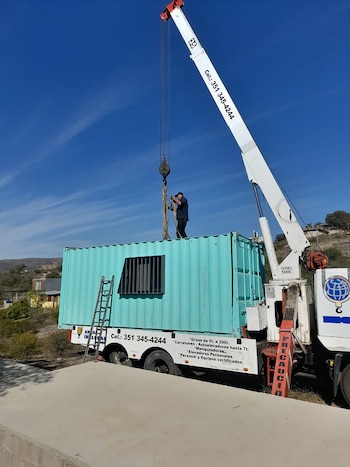 Grúa blanca con brazo extendido levantando un contenedor de envío azul verdoso. Un hombre sobre el contenedor lo guía. Cielo azul claro de fondo