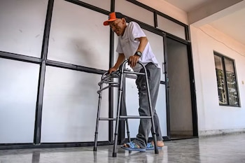 Hombre con gorra, camiseta blanca, pantalón gris, reloj y sandalias azules camina con un andador metálico en un pasillo interior