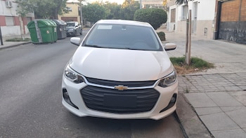 Vista frontal de un automóvil Chevrolet Onix Plus blanco estacionado en una calle urbana. Se observan faros, parrilla, logotipo de Chevrolet, acera y árboles