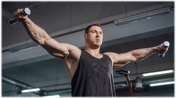 Vista frontal de un hombre musculoso con camiseta negra, levantando dos mancuernas pequeñas a los lados, con los brazos extendidos en un gimnasio.