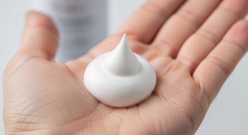 Mousse capilar blanco en la palma de una mano con un envase genérico desenfocado al fondo.
