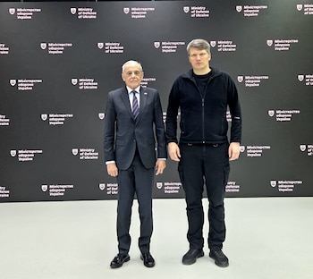 Dos hombres están de pie frente a un fondo negro con logotipos repetidos del Ministerio de Defensa de Ucrania en ucraniano e inglés