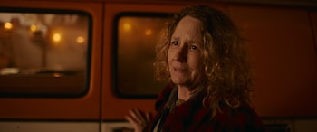 Primer plano de Melissa Leo, una mujer de mediana edad con cabello rizado y rubio, vistiendo una camisa de cuadros rojos y negros, mirando hacia la derecha. Fondo naranja borroso con luces cálidas