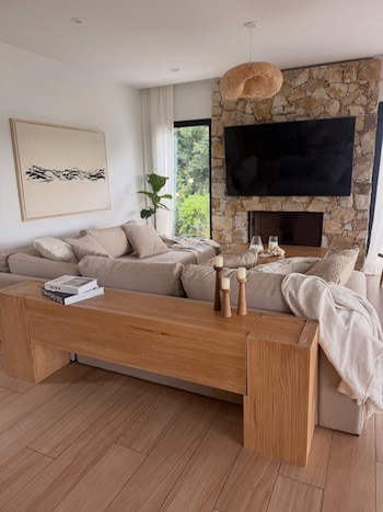 Salón moderno con sofá seccional beige, mesa de centro de madera clara, chimenea de piedra, televisor grande, pintura abstracta y lámpara colgante de mimbre