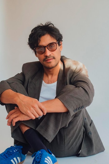 Un hombre de tez oscura, bigote y gafas de sol, vestido con un blazer gris y camiseta blanca, está sentado con zapatillas azules sobre un fondo blanco
