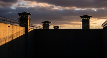 Muros de concreto altos con alambre de púas, tres torres de vigilancia, un cielo nublado y luz del atardecer iluminando una parte del muro.