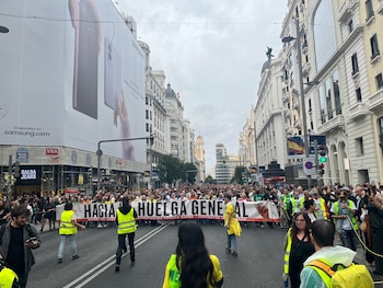 Una multitud de personas camina por una calle flanqueada por edificios y sostiene una pancarta blanca con la frase Hacia la huelga general