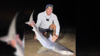Un hombre arrodillado en el agua sostiene un tiburón tigre. Viste gorra, linterna frontal y una cámara al cuello. El fondo es oscuro