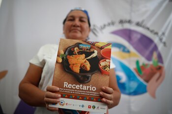 Una mujer sonriente con gorro y delantal blanco con un logo de comida, sostiene una bandeja negra con tres platos de comida. Al fondo, personas y pancartas