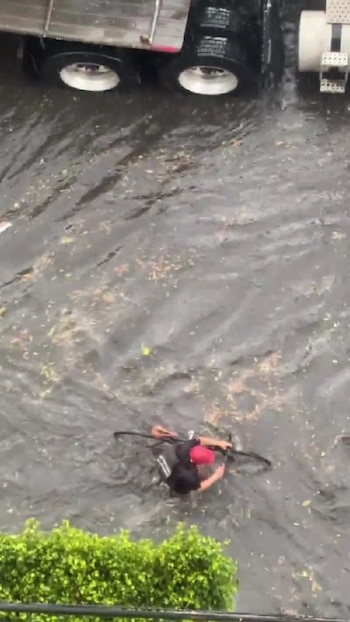 Vista aérea de un ciclista con gorra roja empujando una bicicleta en una calle con agua estancada, un camión y vegetación verde