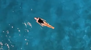 Una persona con traje de baño negro flota de espaldas en agua azul. Hay reflejos de luz sobre la superficie del agua