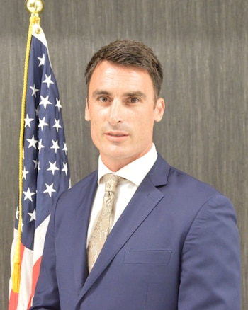 Un hombre vestidocon traje azul y corbata beige posa de frente junto a la bandera de Estados Unidos sobre un fondo gris