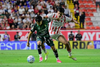 Dos futbolistas, uno con uniforme verde y otro con camiseta rojiblanca, disputan un balón en una cancha de césped con público en las gradas