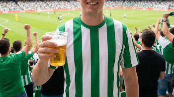 Primer plano de un hombre sonriente con camiseta a rayas verdes y blancas, sosteniendo una cerveza en un vaso transparente en un estadio de fútbol.