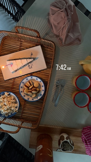 Vista aérea de um prato de bolo com velas, alfajores, pipoca, três xícaras, garfo e esposa, 7h42