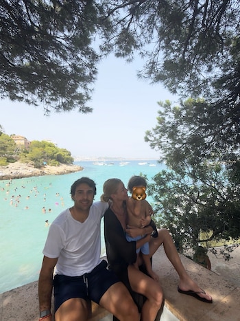 Un hombre, una mujer y un niño sentados en un muro de piedra. Detrás, una cala con bañistas, barcos en el agua, árboles y edificios en la costa
