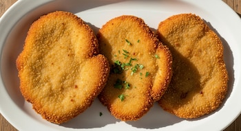 Vista aérea de tres milanesas de ternera doradas y empanizadas, servidas en un plato blanco ovalado sobre una superficie de madera, con perejil fresco en una.