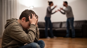 Un niño sentado en el suelo con las manos en la cabeza. Detrás, dos adultos difuminados gesticulan, un sofá en el fondo y una lámpara de techo.