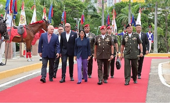 Un grupo de dirigentes políticos y militares uniformados camina sobre una alfombra roja al aire libre, con caballos y banderas de fondo
