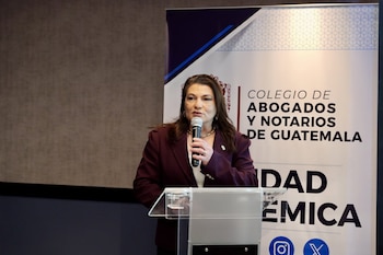 Una mujer habla en un micrófono en un podio transparente. Viste un blazer color vino y un collar de perlas. Detrás, un estandarte dice "Colegio de Abogados y Notarios de Guatemala"