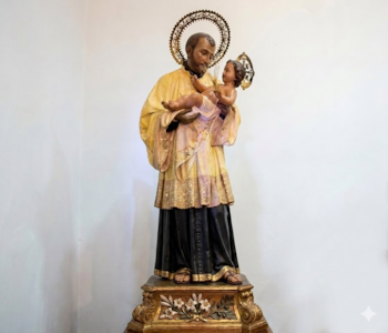 Escultura de San José, con barba y túnica, que sostiene al Niño Jesús. Ambas figuras tienen halo dorado. La base es un pedestal de madera tallada