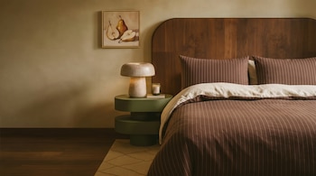 Cama con cabecero de madera y edredón a rayas marrones, mesita de noche verde con lámpara beige, y pintura de peras en pared color tierra.