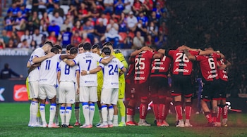 Dos grupos de futbolistas vistos de espalda abrazados en el campo, unos con uniforme blanco con azul y otros con uniforme rojo con negro.