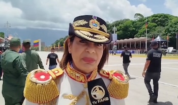 Primer plano de una mujer militar con uniforme de gala y gorra con escudo, frente a un micrófono de TV FANB en una explanada