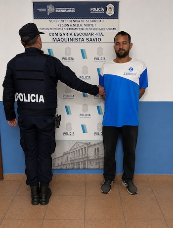 Un policía de espaldas con uniforme azul sujeta el brazo de un hombre con camiseta bicolor y ojos cerrados. Al fondo, un banner de la Comisaría Maquinista Savio