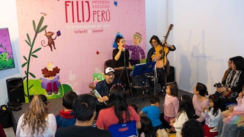 Tres artistas interpretan música y leen ante niños y adultos sentados en el suelo frente a un panel rosado con ilustraciones