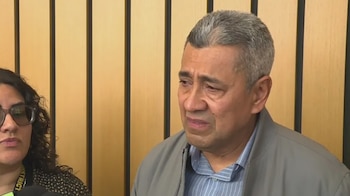 Un hombre de mediana edad, con cabello gris, rostro compungido y chaqueta gris, junto a una mujer parcialmente visible con gafas
