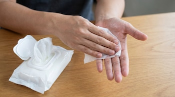 Primer plano de unas manos adultas limpiándose con una toallita húmeda blanca. Un paquete de toallitas húmedas está al lado sobre una mesa de madera clara.