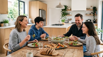 Cuatro personas sentadas a una mesa de madera comen ensalada, vegetales y pollo servidos en platos en el interior de una cocina.
