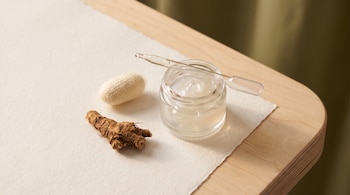 Raíz vegetal seca marrón, capullo de gusano de seda blanco, frasco de gel translúcido con pipeta de vidrio sobre papel y mesa de madera.