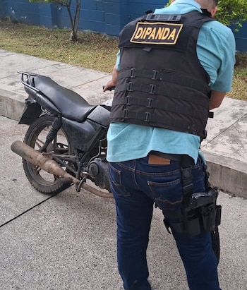 Vista trasera de un oficial con chaleco que dice DIPANDA, camisa celeste, pantalón azul y funda de arma en la pierna, junto a una motocicleta negra