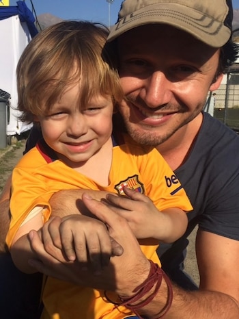 Un hombre adulto con gorra y un niño rubio con camiseta del FC Barcelona posan sonrientes, con el hombre abrazando al niño, en un ambiente exterior soleado
