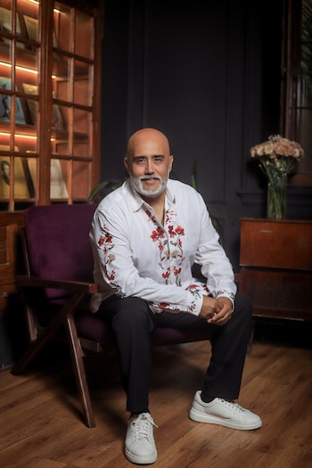 Un hombre con barba canosa y camisa blanca bordada está sentado en un sillón junto a una vitrina iluminada y un florero