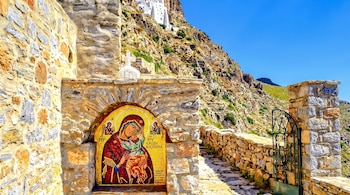 Santuario de piedra con un mosaico de la Virgen María y el Niño Jesús en un camino de montaña con muros de piedra y una verja de hierro.
