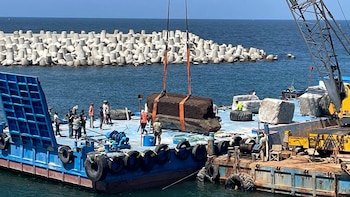 Una grúa levanta un gran bloque rectangular oscuro con correas naranjas de una barcaza azul, con trabajadores y un rompeolas de hormigón en el fondo marino
