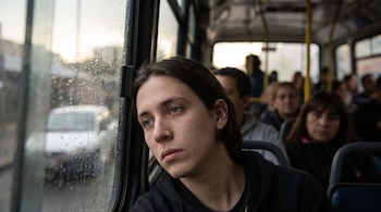 Un joven con cabello largo y mirada pensativa mira por la ventana de un colectivo llena de gotas de lluvia. Otros pasajeros están sentados detrás.