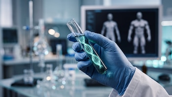 Mano enguantada sostiene tubo de ensayo con hélice de ADN. Fondo con material de laboratorio y monitor con imágenes de cuerpos humanos.
