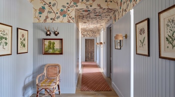 Pasillo con friso de madera azul, papel pintado floral en paredes y techo, una silla de mimbre, cuadros colgados y una alfombra alargada.