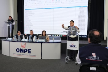 Un hombre sostiene una bolilla numerada de una máquina de sorteo frente a una pantalla con un listado de partidos políticos. Hay varias personas observando