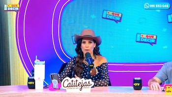 Mujer con sombrero vaquero sentada en una mesa frente a un micrófono en un set de televisión con fondo azul y morado, vasos, un teléfono móvil y un letrero