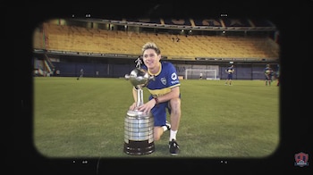 Niall Horan, con camiseta de Boca Juniors, arrodillado en un campo de fútbol, sostiene un gran trofeo. Al fondo, gradas de un estadio con asientos amarillos
