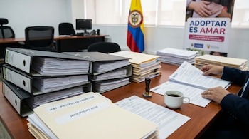 Un escritorio con pilas de documentos, carpetas etiquetadas "PROCESO DE ADOPCIÓN" y una taza de café. Unas manos revisan papeles. Al fondo, la bandera de Colombia y un cartel que dice "ADOPTAR ES AMAR".